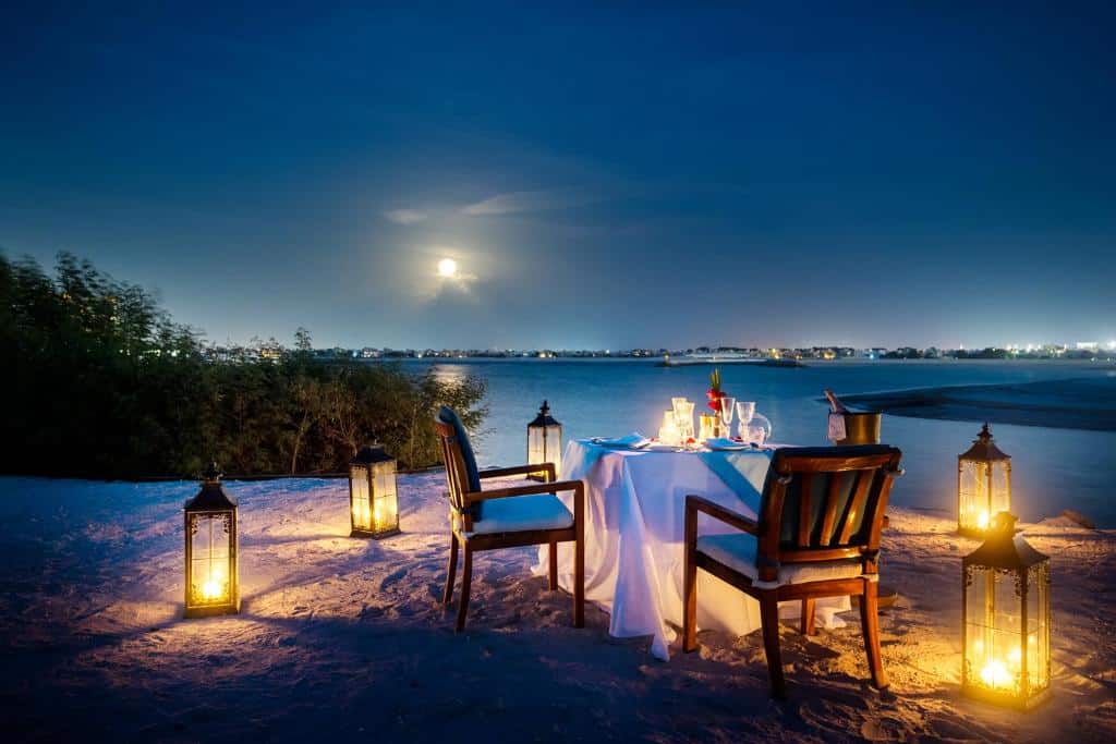 Praia do The Ritz-Carlton Ras Al Khaimah, Al Hamra Beach com uma mesa quadrada com velas e taças, há duas cadeiras de madeira, com alguns lampiões ao redor, para representar hotéis de luxo em Ras Al Khaimah