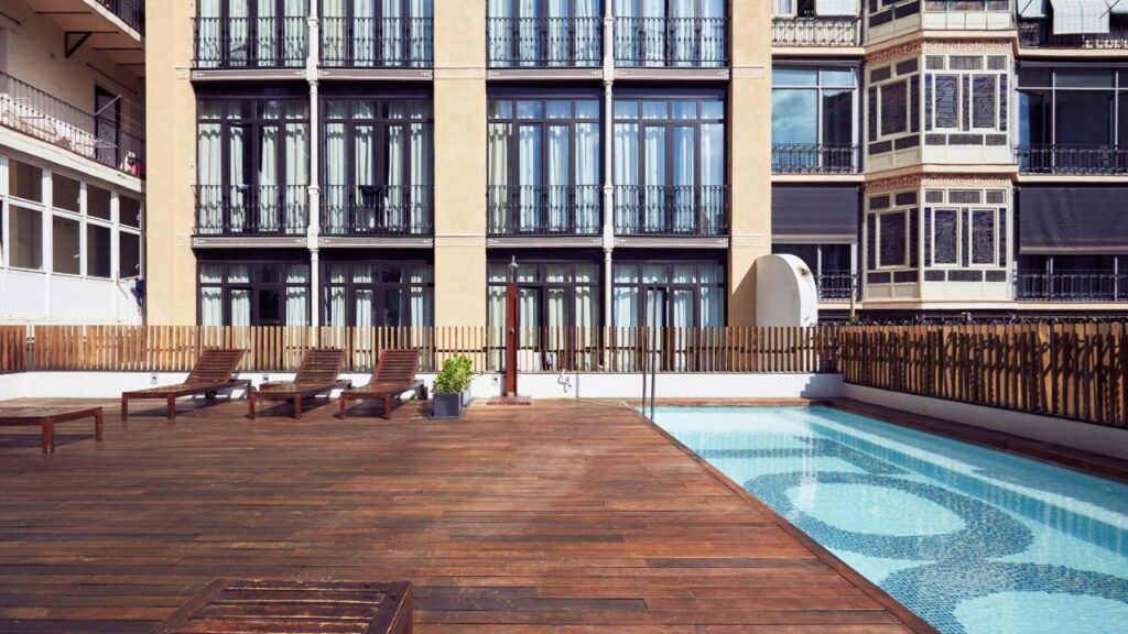 Área da piscina do TOC Hostel Barcelona, uma das recomendações de hostels em Barcelona. Há espreguiçadeiras de madeira com mesa ao lado esquerdo, e a piscina está do lado direito. Ao fundo há fachadas de prédios.