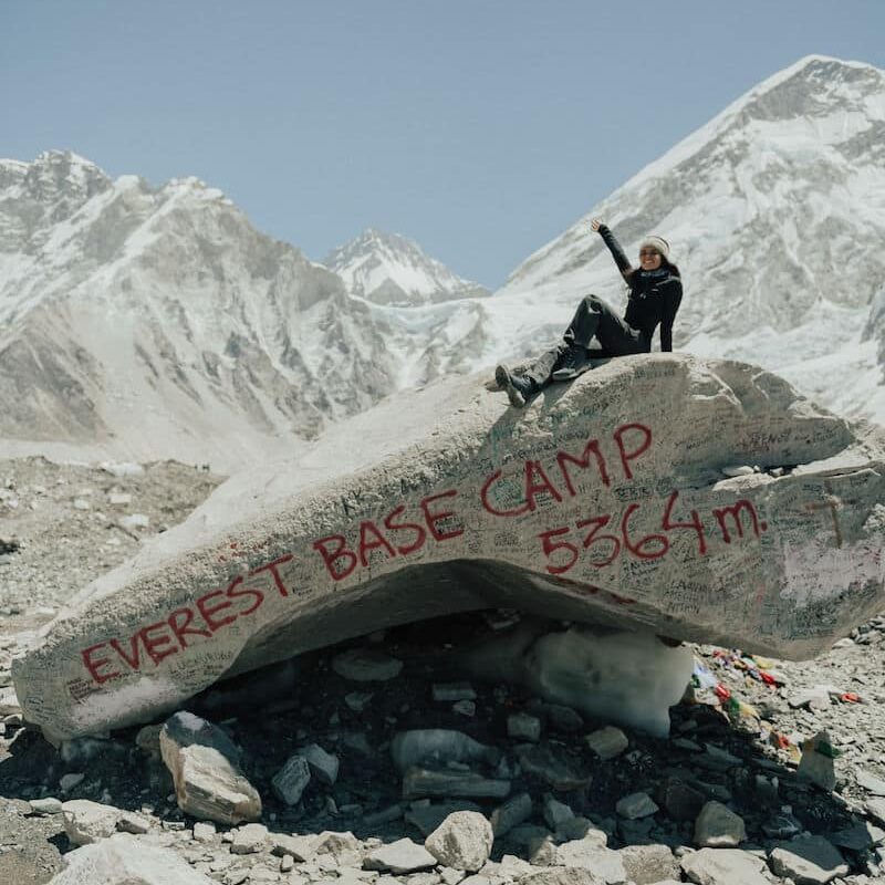 Mulher em cima de uma pedra grande com uma das mãos levantadas com montanhas com gelo atrás. Está de dia, com sol e está escrito de vermelho na pedra "Everest Base Camp 5364m". Foto para ilustrar o post Acampamento Base do Everest
