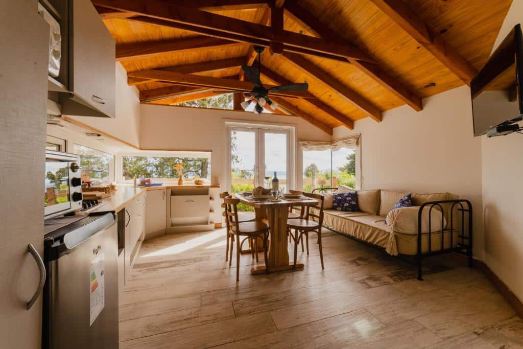 Sala de um aluguel de temporada em Bariloche com sofá, mesa ao meio com quatro cadeiras e janelas de vidro com vista para a natureza. Na parede do lado esquerdo há uma cozinha compacta, com frigobar, forno pequeno, pia, cooktop, e balcão com janelas acima e vista para a natureza