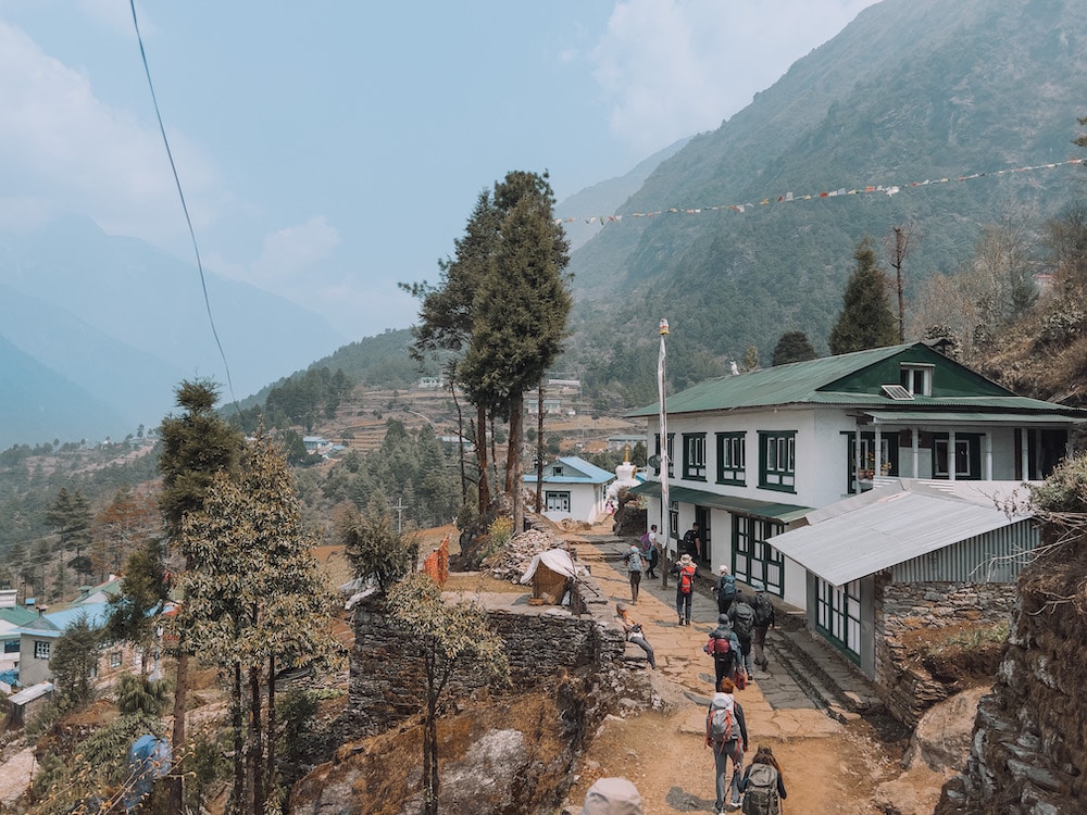 Vilarejo com pessoas andando em uma rua estreita, casas no lado direito e muita natureza nas redondezas. Foto para ilustrar o post acampamento base do Everest