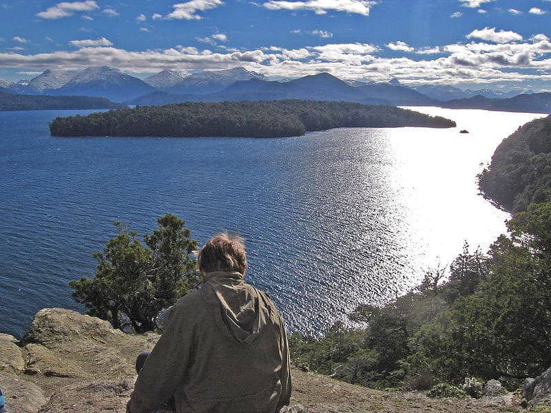 Mulher sentada em uma rocha, de costas, olhando para uma paisagem composta por um lago cristalino com natureza em volta. Está de dia e o céu tem poucas núvens