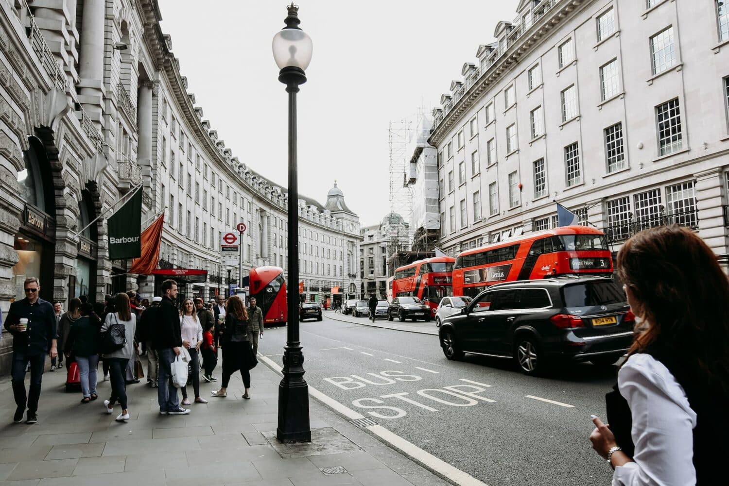 Uma rua movimentada de Londres com ônibus vermelhos e carros passando, há prédios dos dois lados e pessoas caminhando, para representar o aluguel de carro no Aeroporto de Londres