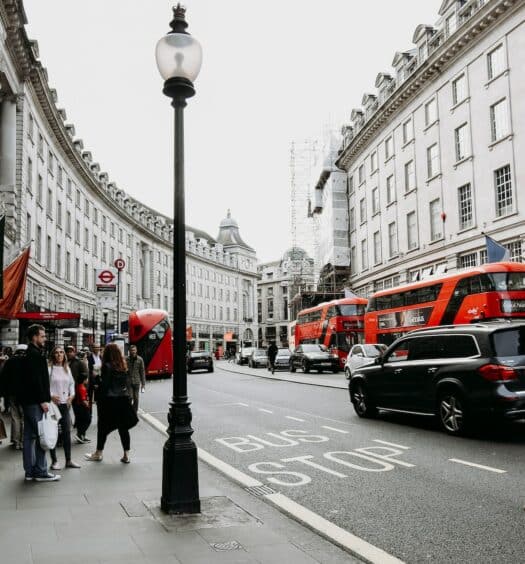 Uma rua movimentada de Londres com ônibus vermelhos e carros passando, há prédios dos dois lados e pessoas caminhando, para representar o aluguel de carro no Aeroporto de Londres