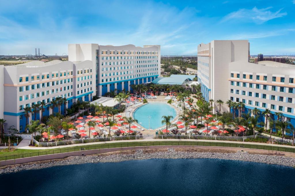 vista do Universal's Endless Summer Resort - Surfside Inn and Suites, um dos hotéis da Universal em Orlando, com uma piscina oval no centro com cadeiras de praia ao redor e prédios de ambos os lados em que o topo parecem ondas, há palmeiras e vista de um lago também