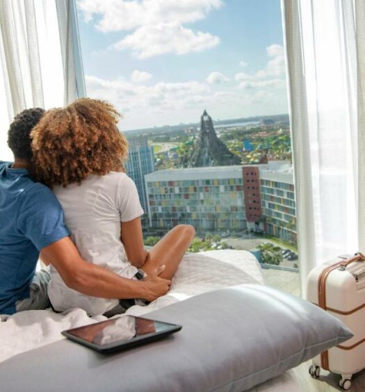 casal de pessoas negras abraçado sentado na cama com vista direta para o Volcano Bay no Universal's Aventura Hotel, um dos hotéis da Universal em Orlando