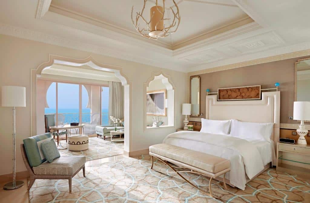 Quarto no Waldorf Astoria Ras Al Khaimah com muito espaço, há dois ambientes, em uma há uma ante sala com sofás e com uma porta de vidro ampla que leva para uma sacada de frente para o mar, do outro ambiente, está o quarto com uma cama de casal grande, um lustre, poltrona, espelho e itens de decoração luxuosos
