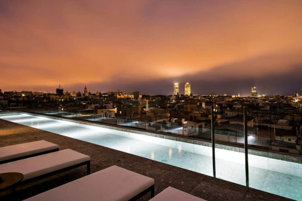 Piscina do Yurbban Passage Hotel & Spa, uma das recomendações de hotéis românticos em Barcelona. A piscina está iluminada e tem espreguiçadeiras brancas por toda a sua extensão. A balaustrada de vidro oferece vista para a cidade de Barcelona. A foto foi tirada à noite.