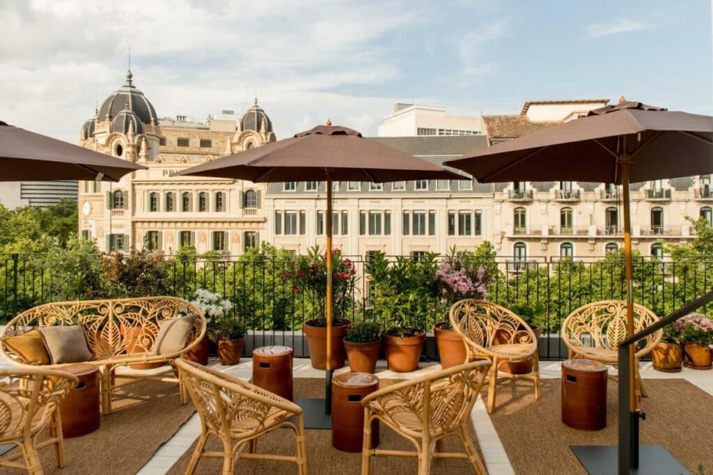 Cobertura do Yurbban Ramblas Boutique Hotel, uma das recomendações de hotéis nas Ramblas em Barcelona. Várias cadeiras com mesas estão dispostas pelo local. Guarda-sóis marrons  vasos de flores estão espalhados por ali também.