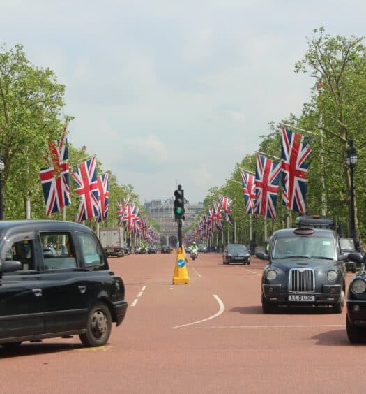Uma rua de Londres com bandeiras penduradas e algumas árvores nas laterais, no centro há diversos carros pretos, para representar aluguel de carro em Londres