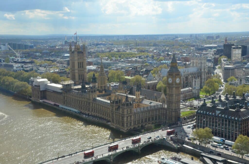 Vista aérea do Palácio de Westminster com destaque para as duas torres principais, sendo uma dela o Big Ben, o rio Tâmisa está na frente do local e há a ponte com diversos ônibus passando e pessoas andando