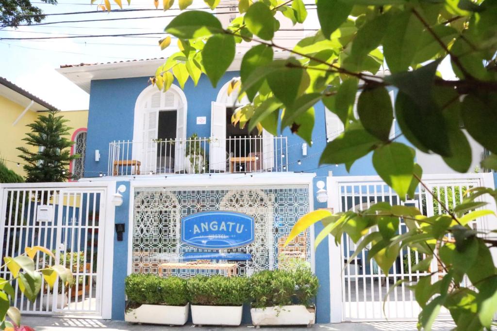 Vista de fora do Angatu Hostel. Sua cor é em um azul médio, com portões e janelas brancos. Uma placa com o nome do hotel está visível no centro. Alguns vasos com plantas decoram o ambiente.