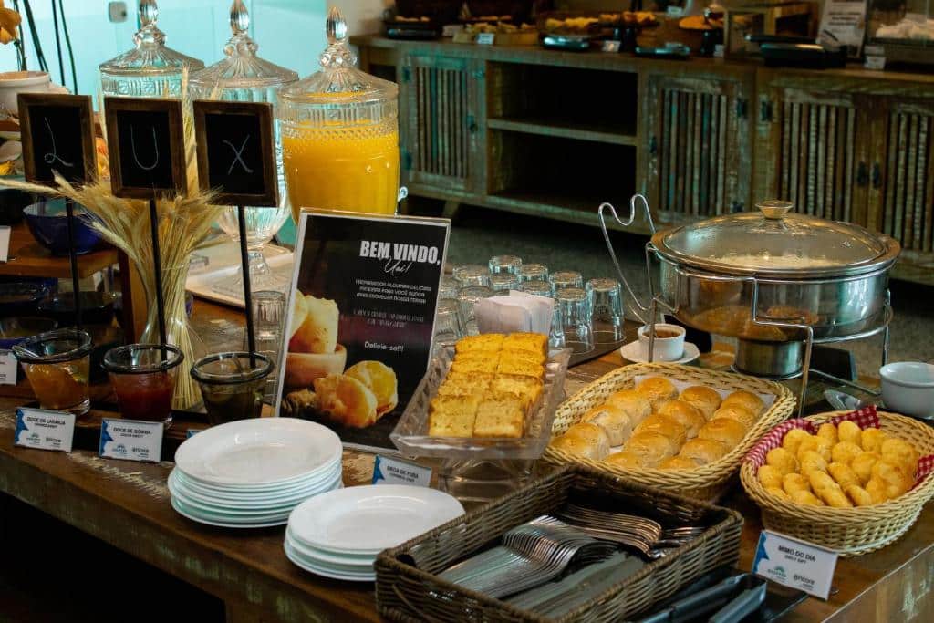 Buffet de café da manhã do Ramada Encore by Wyndham. Há sucos, pães, doces, entre outros alimentos sobre a mesa. É possível ver pratos e talheres.