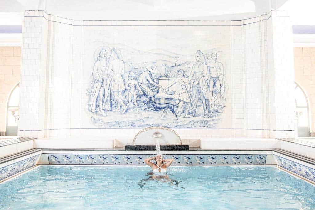 Imagem da piscina do Grande Hotel Termas de Araxá. Há um grande mural com uma pintura clássica/renascentista, os azulejos da piscina são decorados e ela possui um pequeno chafariz, onde há uma mulher molhando seus cabelos embaixo.