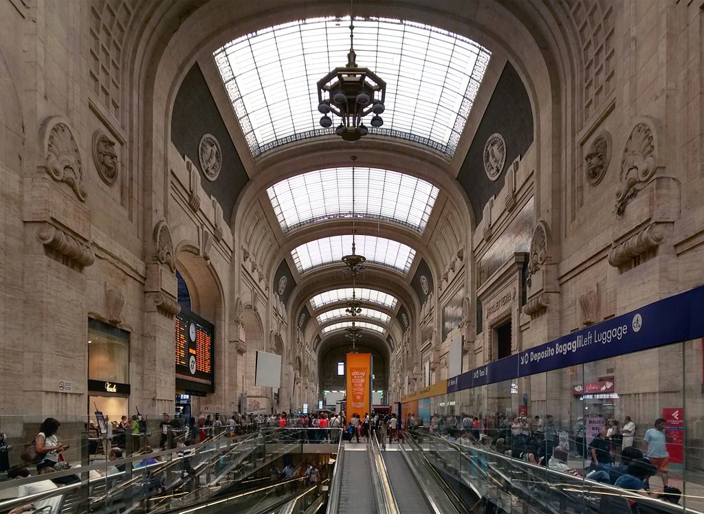 Lado interno da Estação Central, o prédio é muito alto com um teto de vidro, há diversas escadarias, muitas pessoas caminhando e a decoração das paredes lembra o renascentismo