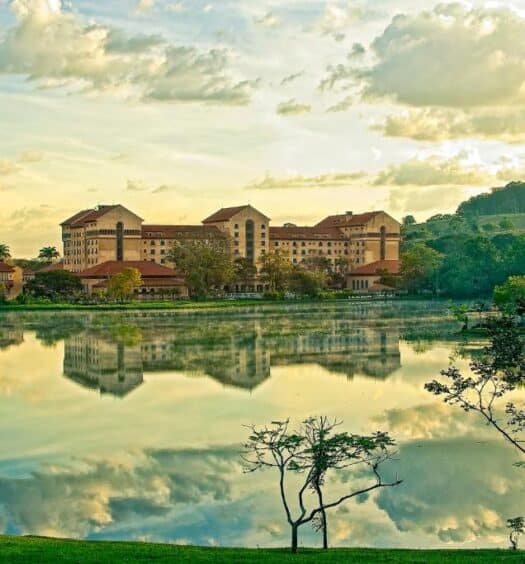 Imagem do hotel Grande Termas de Araxá. O hotel pode ser visto no fundo e no centro da foto, um grande lago está à sua frente, refletindo sua sombra e nuvens no céu. Ao redor do hotel, há várias árvores e espaços verdes. Foto para representar o post de hotéis com piscina aquecida em Minas Gerais.