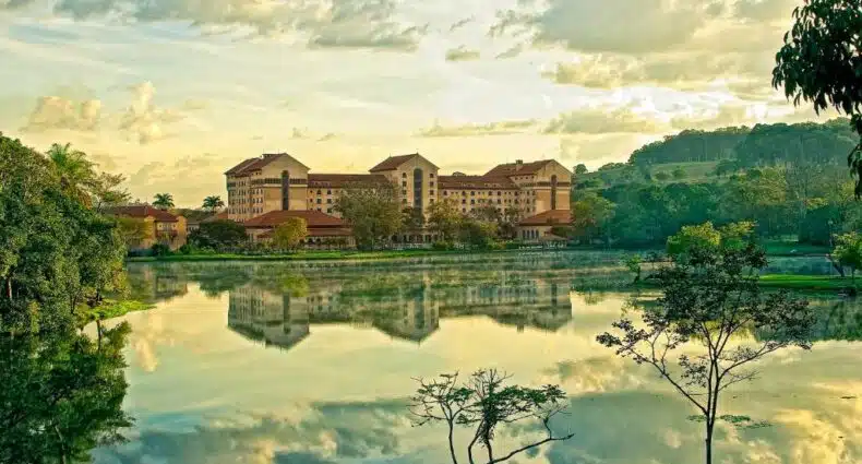 Grande edifício com telhados vermelhos refletido em um lago calmo, cercado por árvores e vegetação sob um céu parcialmente nublado, no Hotel Grande Termas de Araxá, para representar hotéis com piscina aquecida em Minas Gerais.