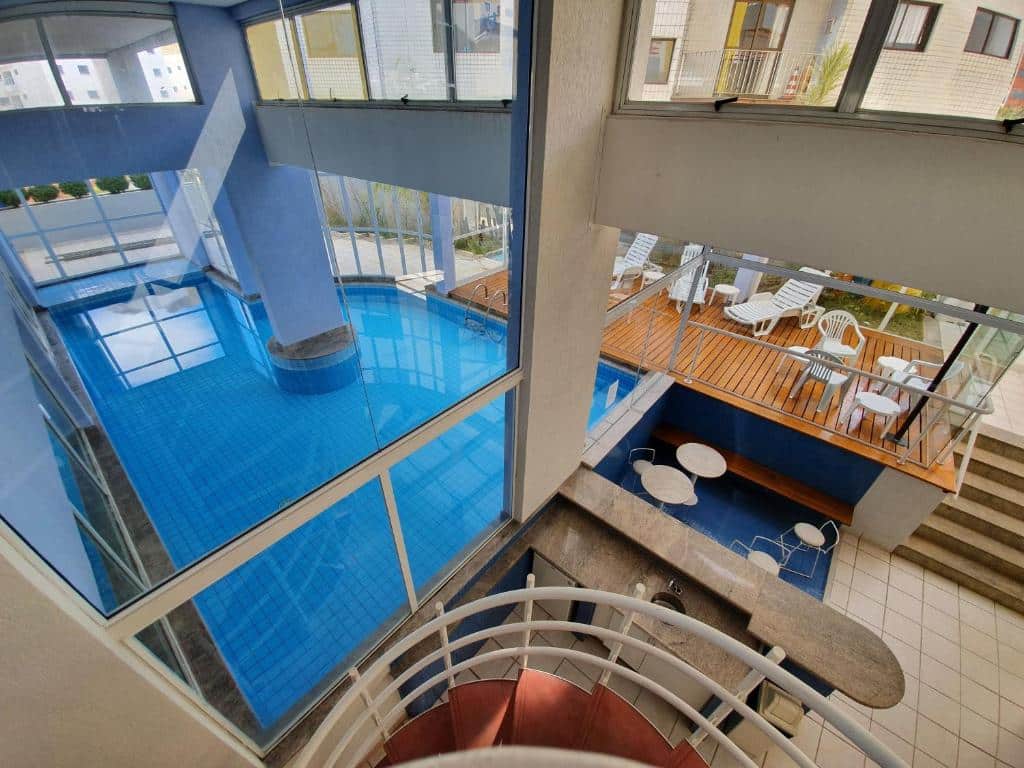 Vista aérea do Luxuoso Flat, imagem para ilustrar o post sobre hotéis com piscina aquecida em Minas Gerais. Temos visão de uma larga piscina, um balcão com mesas e cadeiras e um deck na parte descoberta com espreguiçadeiras e mais cadeiras.
