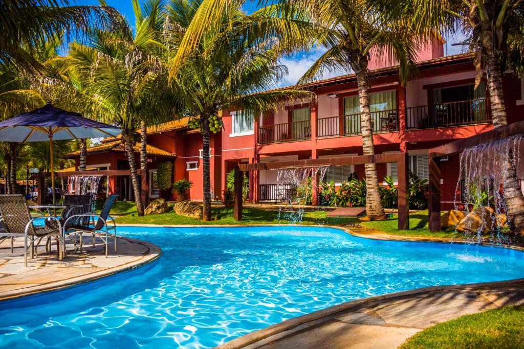 Foto da área externa do Balneário do Lago Hotel, com uma grande piscina, mesas com guarda-sol e cascatas de água. Há bastante espaços com grama e árvores.