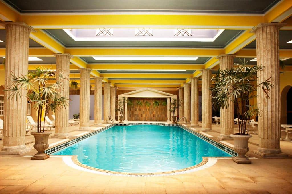 Piscina do Palace Hotel - Poços de Caldas. O ambiente tem um estilo clássico/renascentista, várias colunas estão em volta da piscina. O teto possui claraboias. Há espreguiçadeiras e vasos com plantas pelo ambiente.