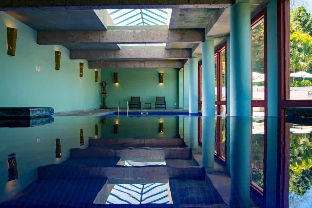 Piscina interna e coberta do hotel Hotel Solar do Rosário. Uma claraboia está no teto. As paredes são verde-água, e algumas colunas estão posicionadas perto das janelas. Duas espreguiçadeiras estão no fundo da imagem.