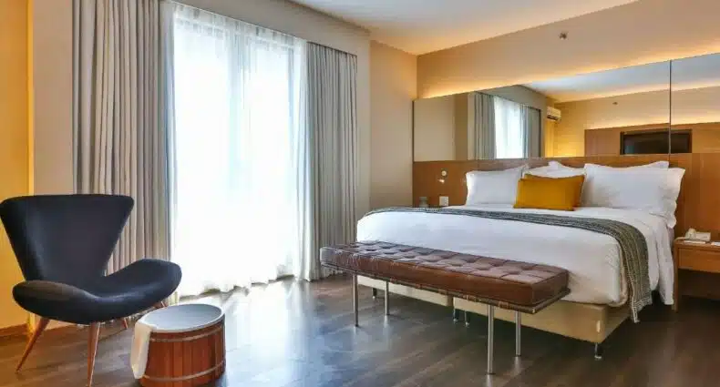 Suíte no Radisson Blu São Paulo. A cama é espaçosa, os lençóis e travesseiros são brancos. Uma almofada no centro é amarela. Há um pequeno sofá-cama de couro marrom no pé da cama. Atrás do móvel, um largo espelho está fixo na parede, tendo uma iluminação de LED atrás do mesmo. Do lado esquerdo da imagem, está a janela do quarto com cortinas claras. Uma poltrona preta está posicionada perto dela. Foto para representar o post sobre hotéis na Faria Lima em SP.