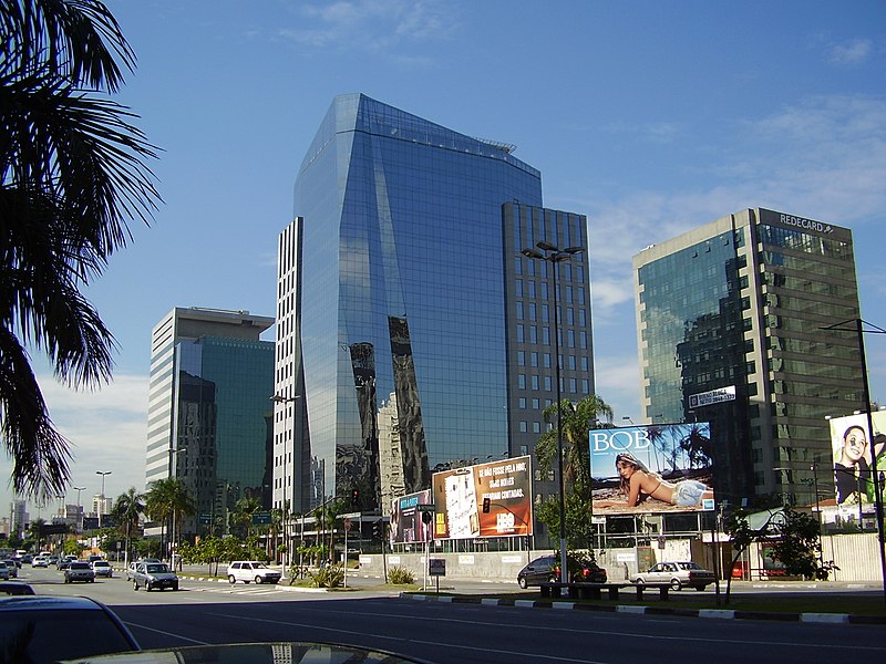 Vista de uma avenida movimentada na Vila Olímpia, com múltiplos outdoors, prédios e arranha-céus. Foto tirada de dia, usada para representar o post sobre hotéis em Vila Olímpia.