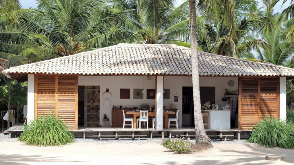 Vista externa da propriedade Abaetetuba Pousada, de localização pé na areia em Milagres, no Alagoas, Atrás da casa há folhagens de coqueiros, na parte aberta é possível ver a mesa do café da manhã, e à frente, depois de um desnível, já começa a praia