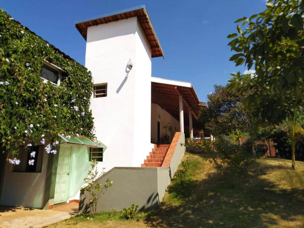 Casa branca com escadas para a parte de cima, uma porta bem no começo da escada e bastante árvores no gramado verde.