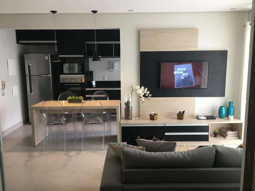 Uma sala com sofá e televisão, atrás a vista para cozinha, com mesa, geladeira, microondas e pia.