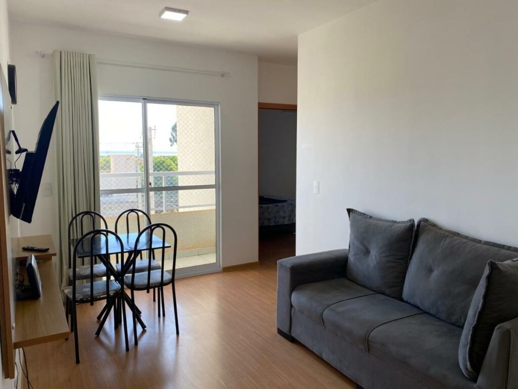Foto do interior do apartamento Apês Palmeira Dourada - Centro de Palmas. É possível ver um sofá, uma mesa pequena e quadrada com quatro cadeiras, televisão, uma porta que leva até a sacada e, ao fundo, uma parte do quarto.