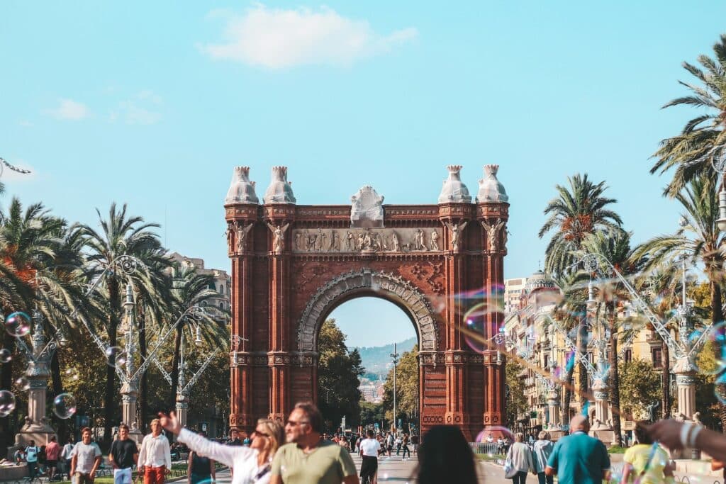 Pessoas caminhando na rua em frente ao Arco do Triunfo em Barcelona. Há palmeiras dos dois lados da ampla rua, e o céu acima está azul. - Foto: Rachel Moore via Unsplash