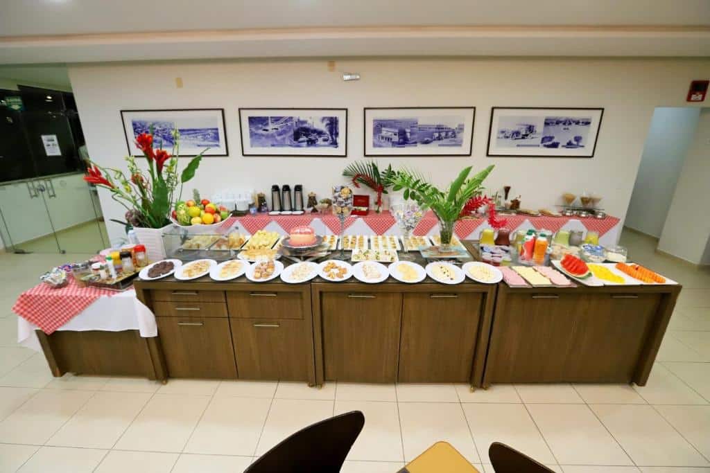 Mesa com café da manhã de hotel com grande variedade de frutas, salgados, sucos e duas plantas decorativas em cima da mesa.