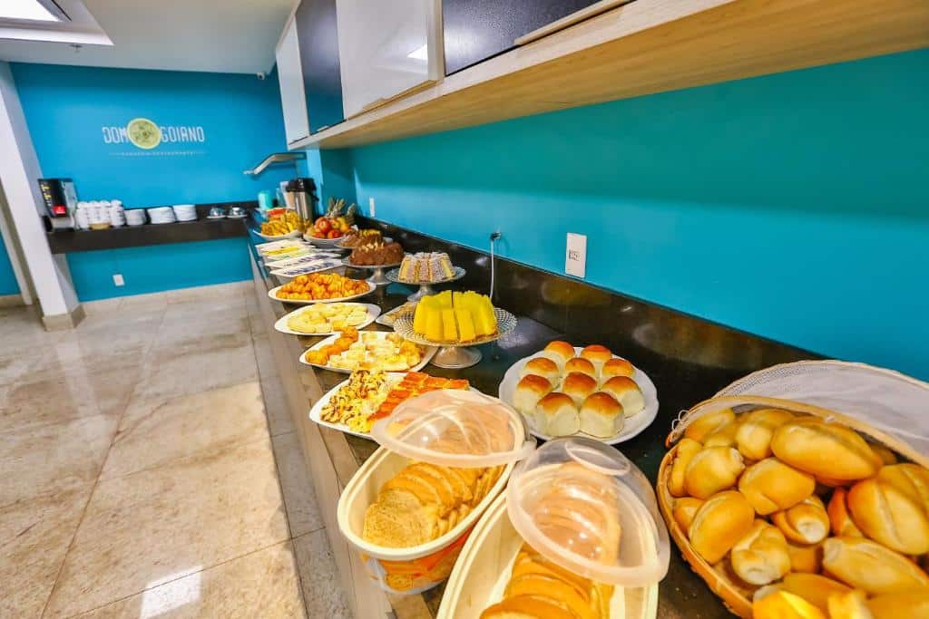 Saguão de hotel com paredes azuis e bancada com café da manhã variado com muitos pães, bolos, salgados e frutas.