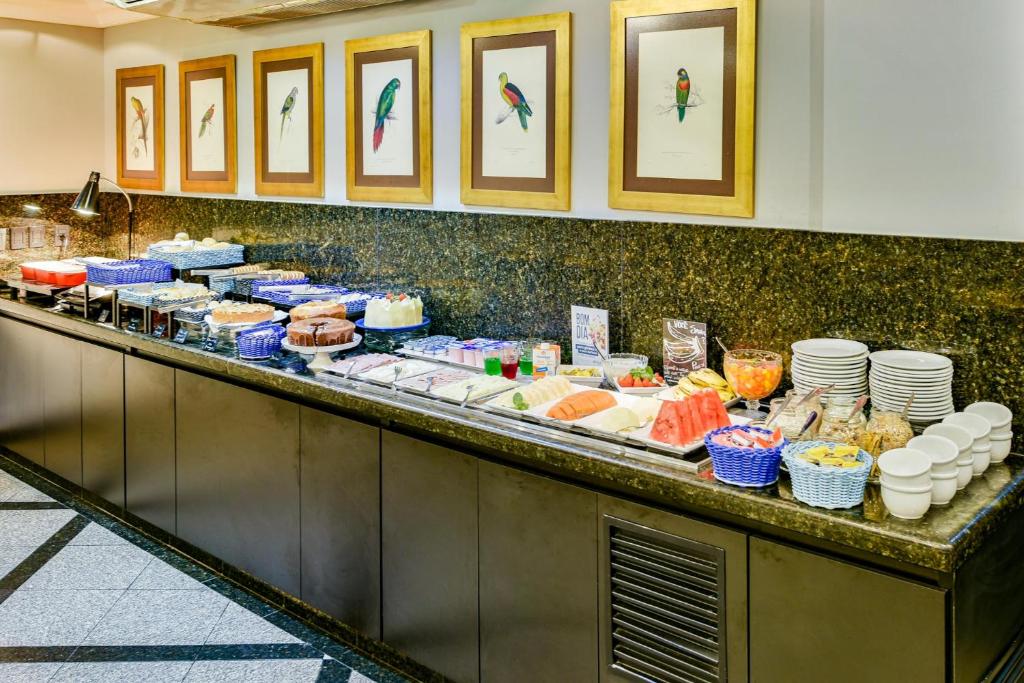 Bancada de hotel com café da manhã variado, bolos, salgados, cestas com pães, frutas e pratos.