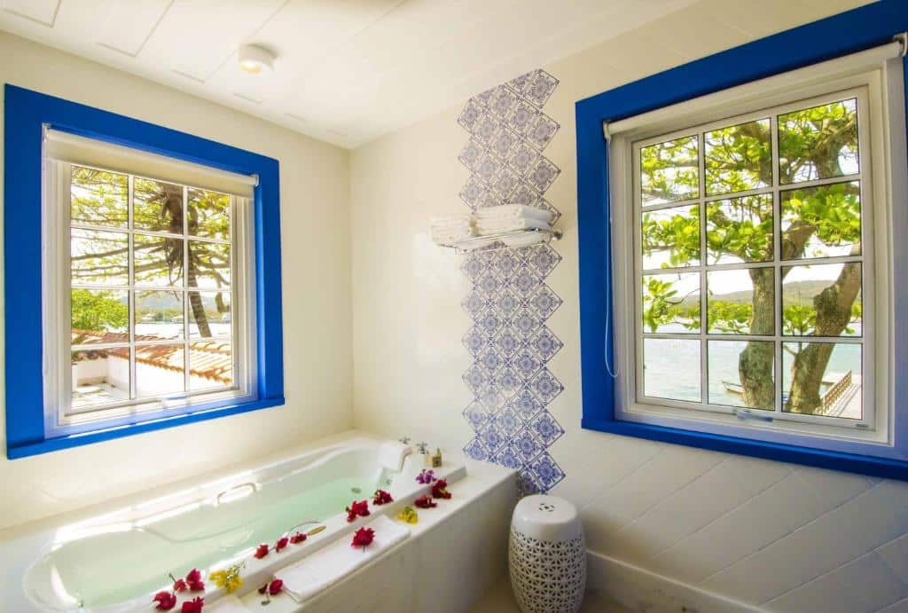 Banheiro de hotel com banheira de hidromassagem, flores vermelhas e janelas com vista para o lago com molduras azuis.