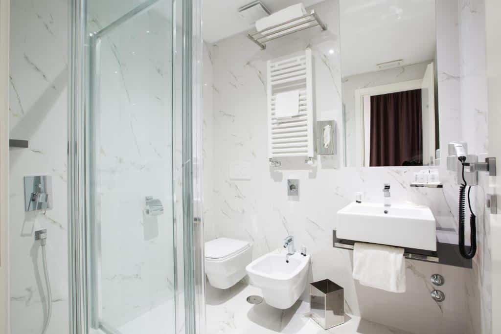 banheiro do Crosti Hotel com box de vidro aberto, chuveirinho, bidê, vaso sanitário mais baixo, pia com espaço abaixo