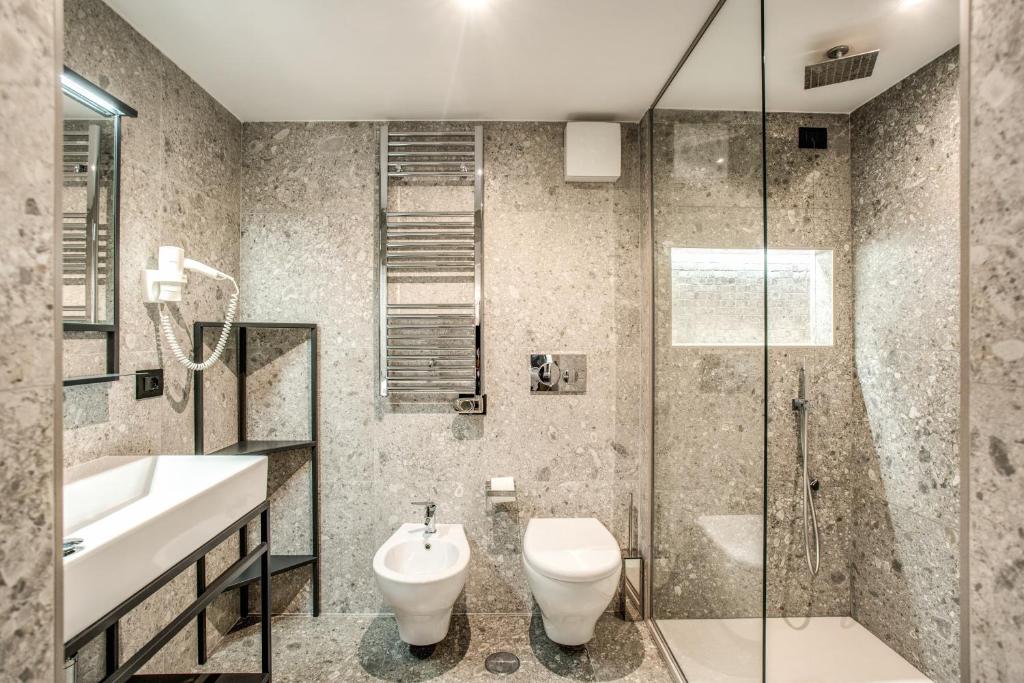 banheiro do FOURHEADS Private Suites, com vaso sanitário e bidê, pia com espaço embaixo, prateleira aberta, box aberto com chuveirinho, todo revestido em pedra