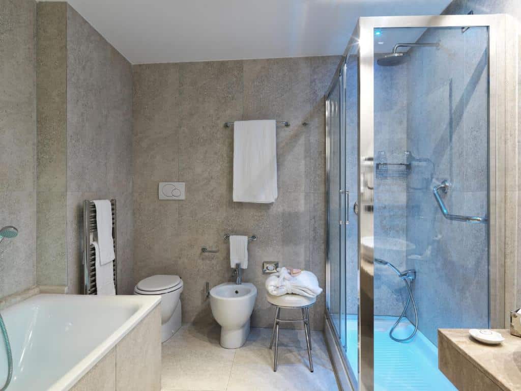 banheiro do Hotel Barocco, um dos hotéis no centro de Roma, com vaso sanitário e bidê, cadeira de banho, box aberto com chuveirinho e barras de apoio