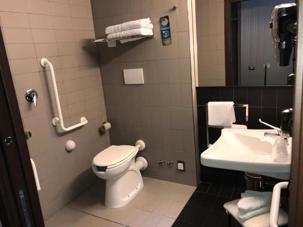 banheiro do Ibis Styles Roma Eur um dos hotéis baratos em Roma, com adaptações para PcDs, sendo elas: pia mais baixa com espaço abaixo, vaso sanitário elevado, barras de apoio e cadeira de banho em um ambiente aberto
