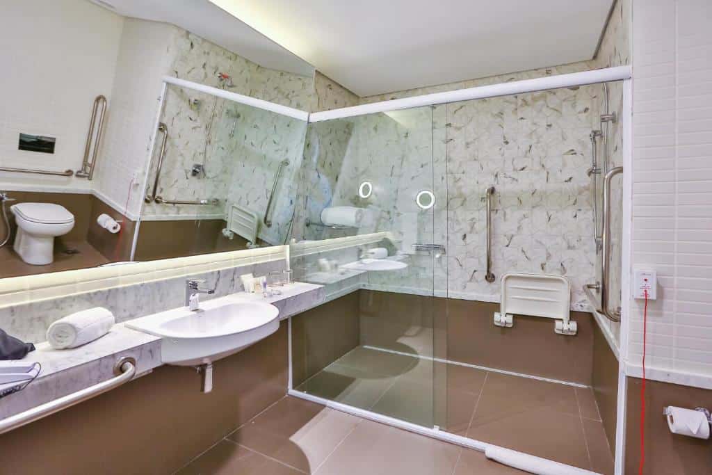 Banheiro com azulejos brancos e marrons, pia mais baixa, barras de apoio do lado de fora e dentro do box para melhor acessibilidade para hóspedes com mobilidade reduzida. Imagem para ilustrar o post hotéis em Goiânia.
