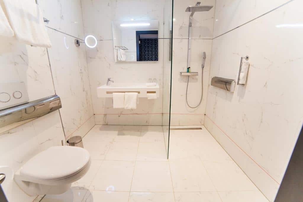 Banheiro adaptado do DoubleTree, em Amsterdam, com chão e paredes de mármore branco, um vidro separando a área do banho e a pia e um vaso branco. O vaso e a pia são baixos e há bastante espaço entre os itens