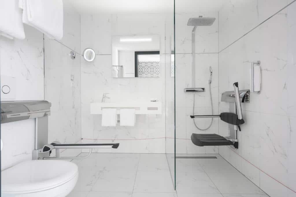 Banheiro com acessibilidade do DoubleTree by Hilton Amsterdam, com áre de banho com cadeira de banho, barras de apoio, e ao lado tem uma pia baixa e a frente um vaso com barra de apoio ao lado