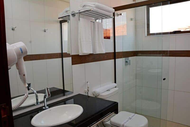 Banheiro de hotel com azulejos brancos, secador de cabelo e box ao lado da privada.
