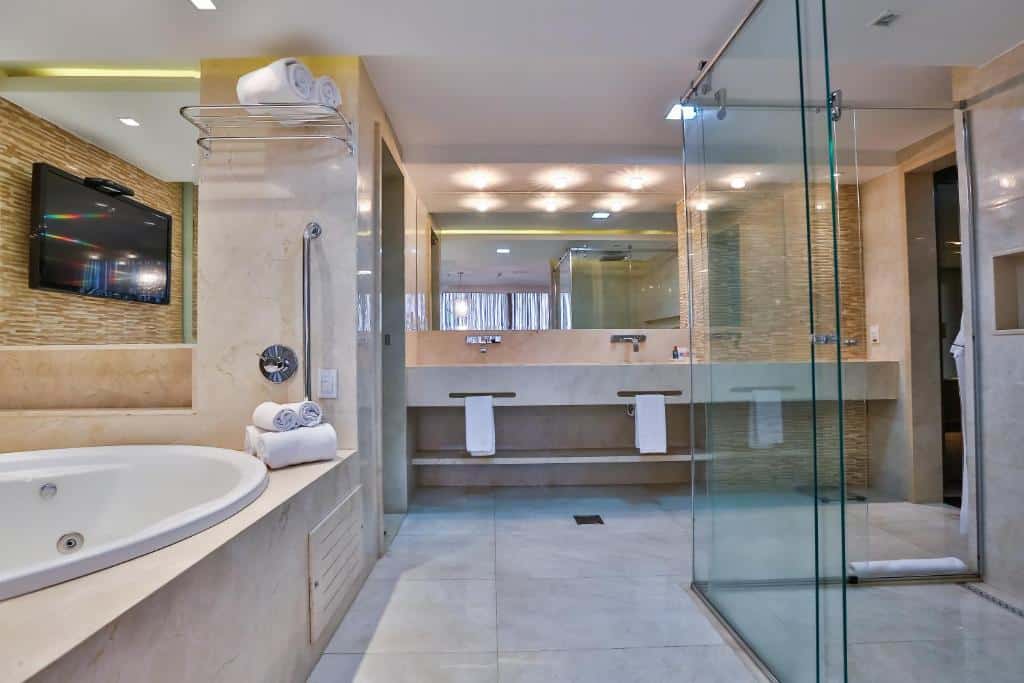 Banheiro de hotel luxuoso com paredes em mármore branco, três pias, um espelho, uma banheira de hidromassagem e um box grande de vidro.