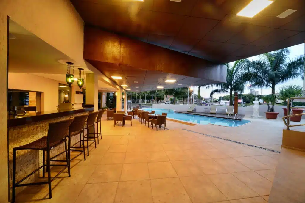 Área externa com uma piscina, árvores e um bar com mesas e cadeiras em volta. Foto para ilustrar post sobre hotéis em Campo Grande.