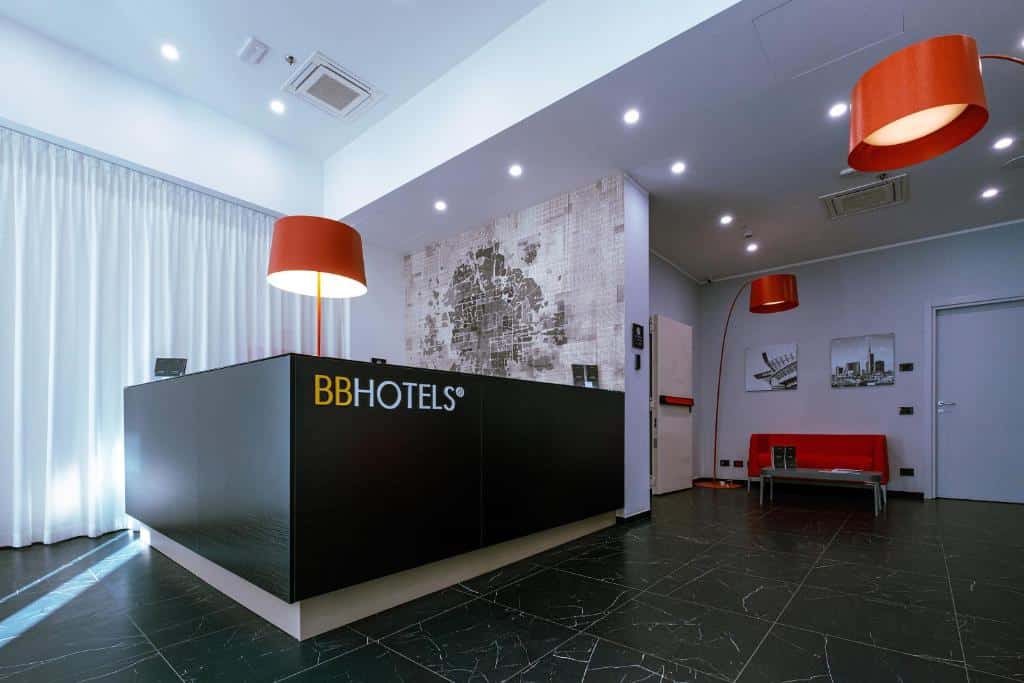 Recepção do BB Hotels Smarthotel Duomo com um balcão preto, chão preto, algumas luminárias vermelhas e um sofá vermelho