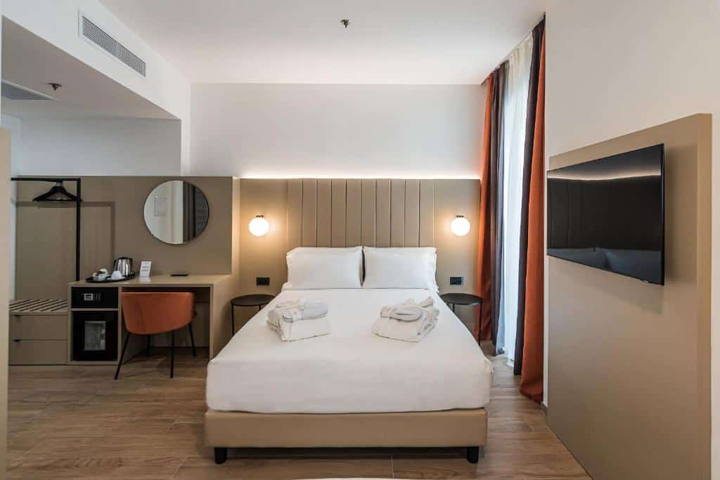 Quarto do BB Hotels Smarthotel Duomo com uma cama de casal, uma janela com cortinas, uma televisão, uma cabeceira com luminárias, uma mesa de escritório com uma cadeira e um pequeno armário de conceito aberto, para presentar hotéis perto do Duomo em Milão