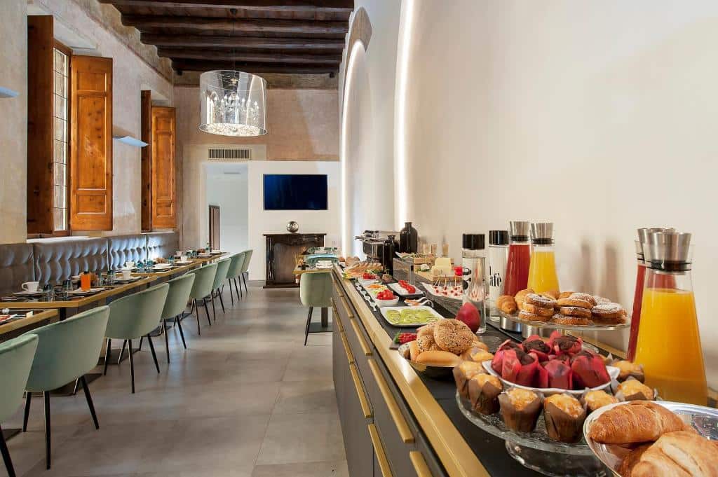 salão do Argentina Residenza Style Hotel com poltronas esverdeadas com mesinhas e um grande lustre acima e um balcão com doces, frutas e sucos