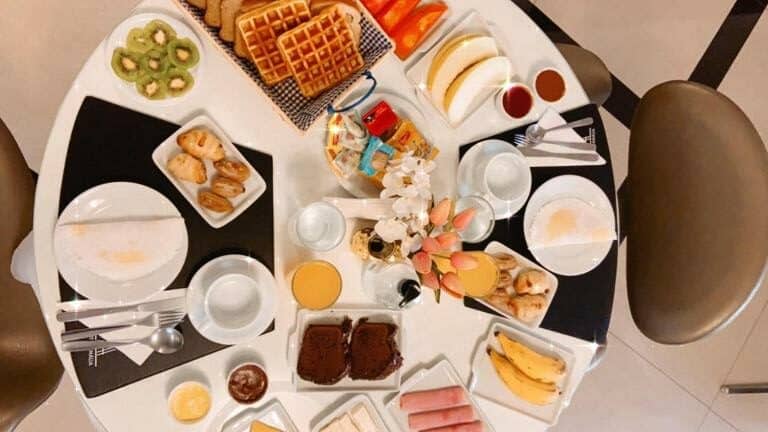 Uma mesa com o café da manhã, com uma grande variedade. Bolos, frutas, pães e frios.